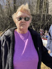 a woman wearing sunglasses and a purple shirt is standing in the woods