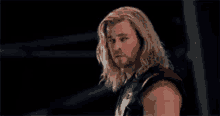 a man with long blonde hair and a beard is standing in a dark room looking at the camera .