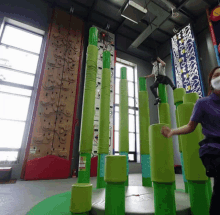 a woman wearing a mask stands in front of a climbing wall while a man climbs on a set of green poles