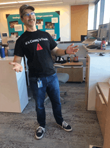 a man wearing a casey 's fault t-shirt stands in an office