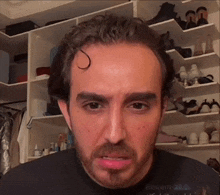 a man with a beard and a black shirt is standing in front of a closet with shoes on shelves