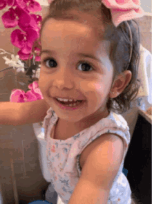 a little girl with a pink bow in her hair is smiling in front of a vase of flowers .