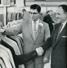 two men shaking hands in front of a rack of suits