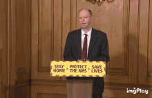 a man stands at a podium holding a sign that says " stay home protect the nhs save lives "