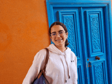 a woman wearing glasses and a white under armour hoodie smiles in front of a blue door