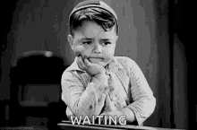 a black and white photo of a little boy sitting at a desk with his hand on his chin and waiting .