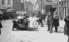 a black and white photo of a car driving down a city street with people standing on the sidewalk .