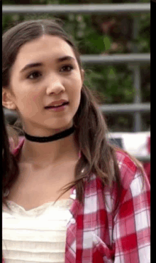 a young girl wearing a choker and a plaid shirt is standing next to a fence .