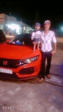 a man is holding a baby while standing next to a red car at night .