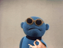 a blue stuffed animal wearing sunglasses is being held by a person 's hand .