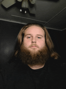 a man with long hair and a beard is standing in front of a ceiling fan