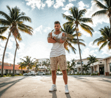 a man in a white tank top and khaki shorts is standing in front of palm trees