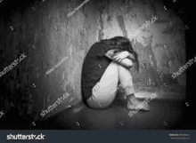 a black and white photo of a woman sitting in a corner of a room with her head down .