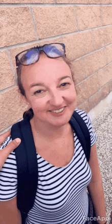 a woman wearing sunglasses and a striped shirt is smiling and standing in front of a brick wall