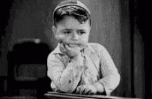 a black and white photo of a little boy sitting at a desk with his hand on his chin .