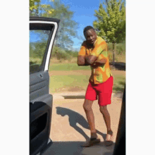 a man in a colorful shirt and red shorts is standing in front of a car with his arms crossed .