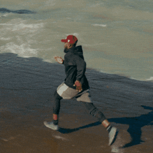 a man wearing a red hat and a black hoodie is running on the beach