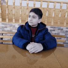 a boy in a blue jacket sits at a table with his hands folded in front of him