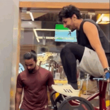 a man in a red shirt is standing next to a man in a black tank top who is riding an exercise bike