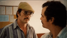 two men are talking to each other in a room . one of the men is wearing glasses and a hat .