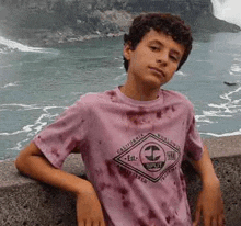 a young boy wearing a pink tie dye t-shirt is leaning against a wall next to the ocean .