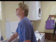 a man in a blue shirt is standing in a laundry room with a purple bag hanging on the wall