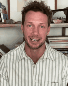 a man with a beard wearing a striped shirt is smiling in front of a bookshelf filled with books .