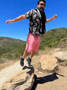 a man in a floral shirt and pink shorts jumps over a rock