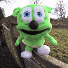 a stuffed green gummy bear with blue eyes is sitting on a wooden fence