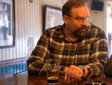 a man in a plaid shirt sits at a table with a glass of beer on it