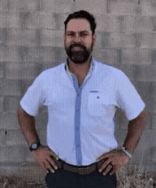 a man with a beard wearing a white shirt is standing with his hands on his hips in front of a brick wall .