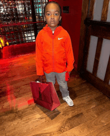 a little boy wearing an orange nike jacket holds a red bag