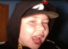 a young boy wearing a baseball cap and a hoodie is sticking his tongue out .