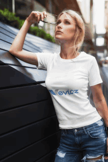 a woman leaning against a wall wearing a white t-shirt that says novioz