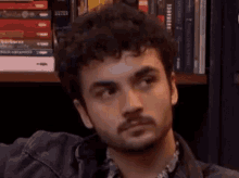 a man with a beard and curly hair is sitting in front of a bookshelf with books on it .