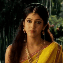 a woman wearing a yellow saree and gold jewelry looks at the camera