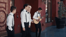 a group of men in tuxedos and suspenders are standing in front of a red building one man is playing a guitar