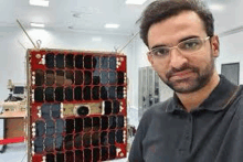 a man wearing glasses is standing in front of a small satellite .