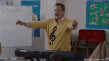 a man wearing a yellow sweater with a treble clef on it stands in front of a white board that says tucker