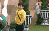 a young boy in a yellow shirt and black pants is standing in front of a potted plant in a garden .