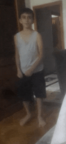 a young boy in a white tank top and black shorts is standing in a room with his hands in his pockets .