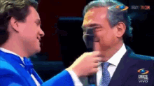 a man in a blue tuxedo is touching another man 's face in front of a screen that says caracol vivo