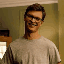 a young man wearing glasses and a grey t-shirt is smiling in a living room .