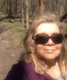 a woman wearing sunglasses and a purple sweater stands in the woods