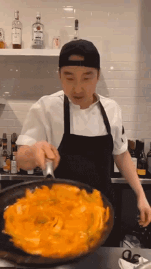 a man in an apron is stirring a pan of food with a bottle of bombay sapphire in the background