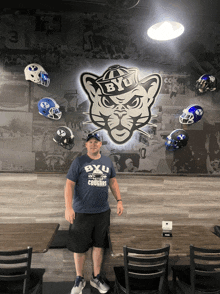 a man wearing a byu shirt stands in front of a wall with football helmets hanging from it