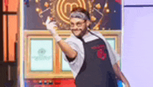 a man in an apron and gloves is standing in front of a wall with a master chef logo on it .