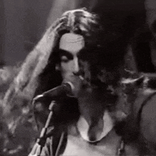 a man with long hair is singing into a microphone while smoking a cigarette .