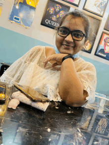 a woman wearing glasses sits at a table with a queen poster on the wall behind her