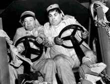 two fat men are sitting next to each other holding steering wheels .